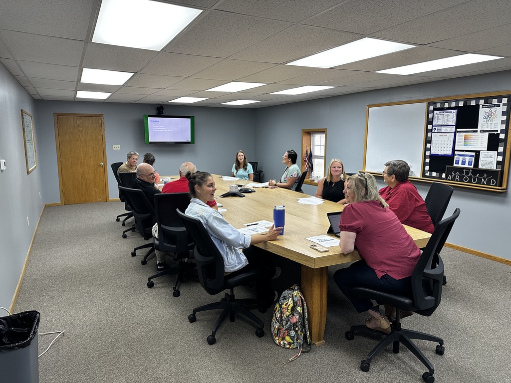 A group of people sitting at a table in a meeting room with a monitor on the wall.