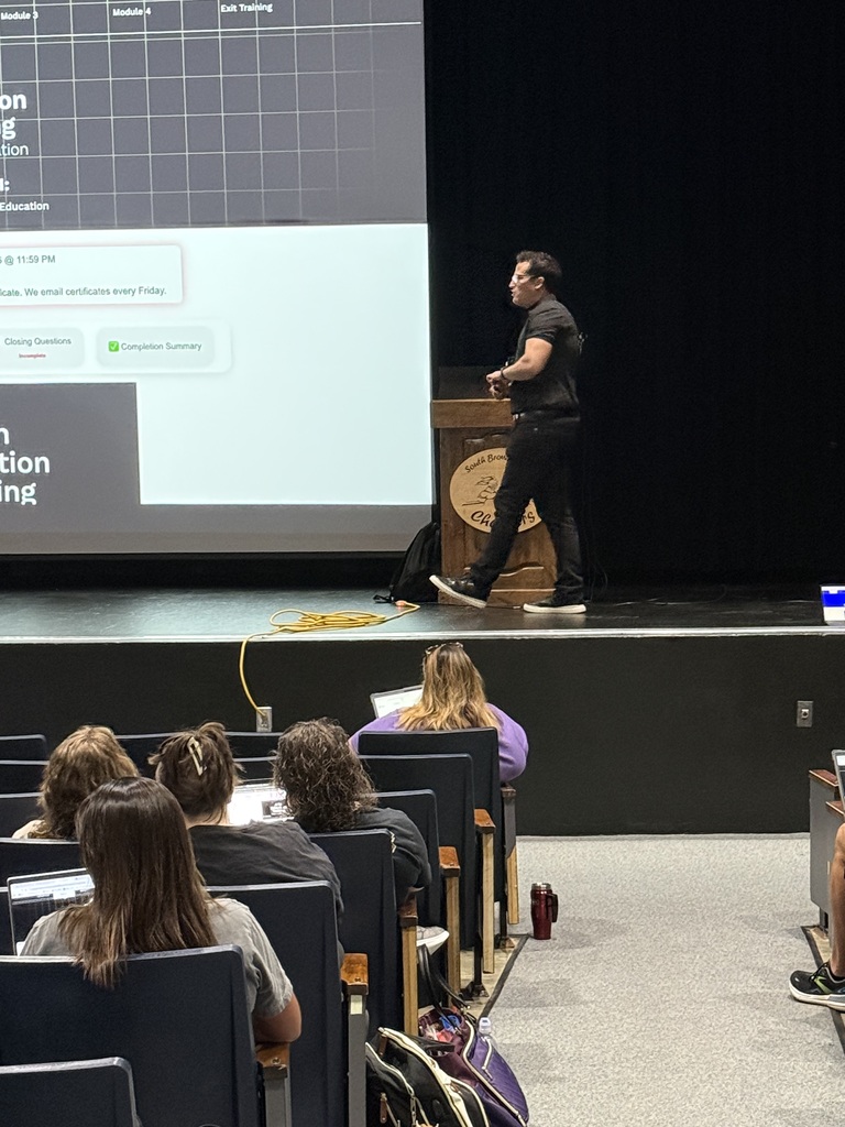 A classroom with a person on stage giving a lecture in front of an audience seated in rows.