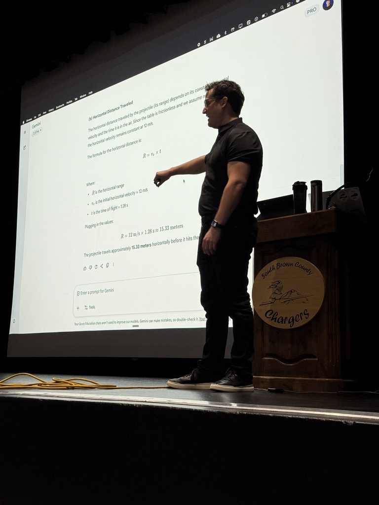 A man on a stage with a large screen behind him, displaying a presentation. He points at the screen with his right hand.