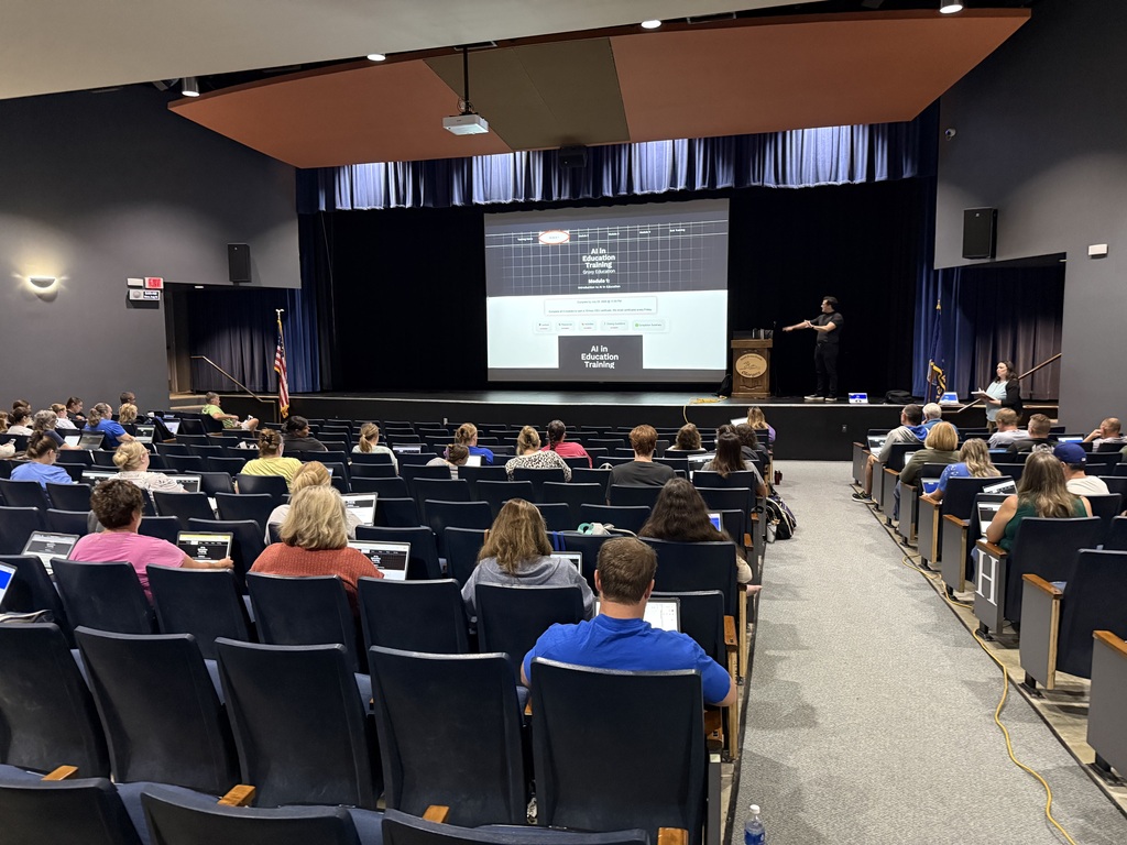An auditorium with rows of chairs, a person presenting at a podium, a projector screen displaying a presentation.