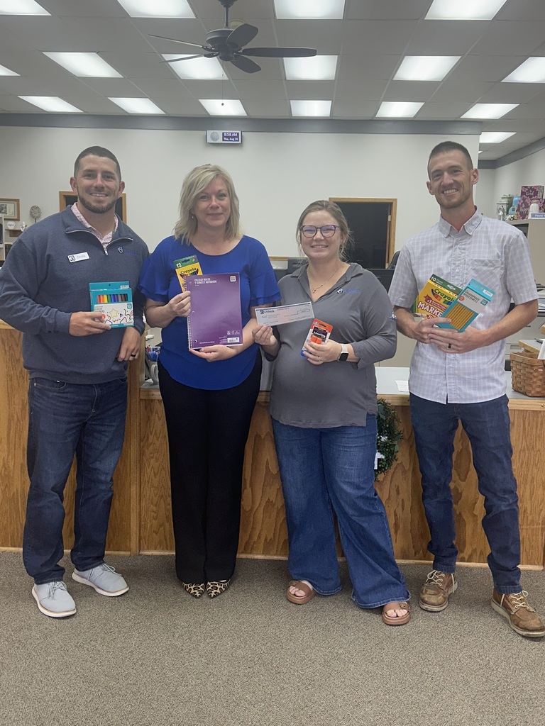 Four people stand together in a room, holding books. They are smiling. A desk with decorations is behind them.