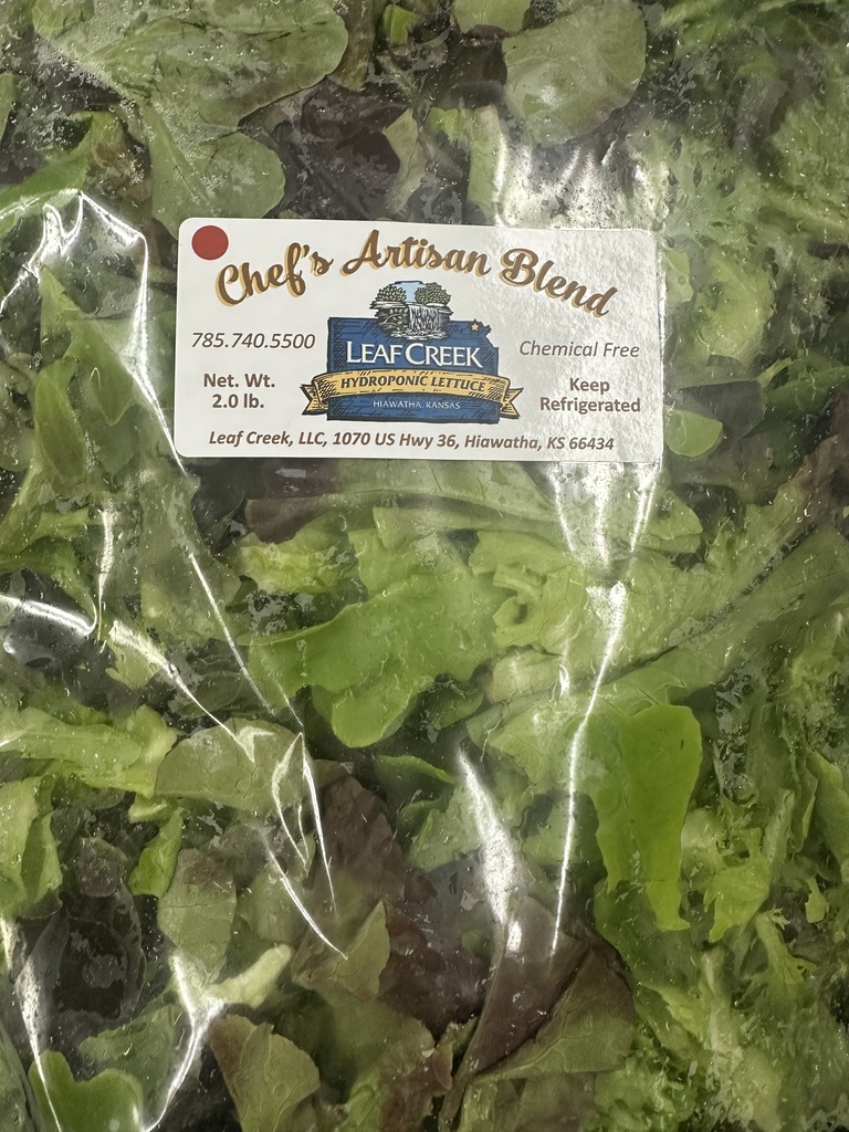 Plastic bag with a white label containing text. Inside the bag is a mix of dark green and light green leafy vegetables.