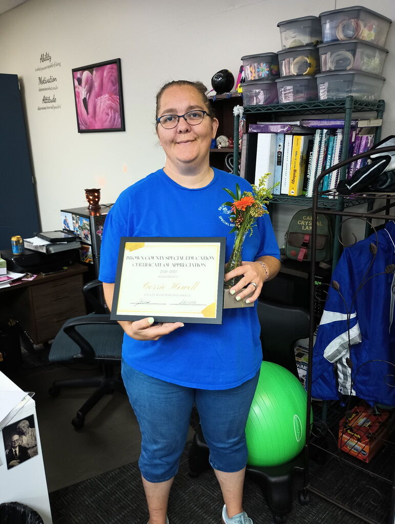 A woman stands in an office, holding a framed certificate and a flower vase. Behind her, a desk has books, a storage box, and a ball.