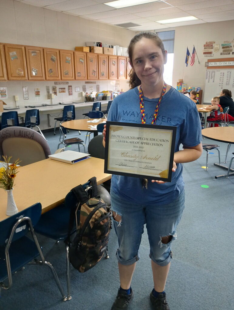 A woman in a blue shirt holds a framed certificate, standing in a classroom. The room has desks, chairs, and shelves.