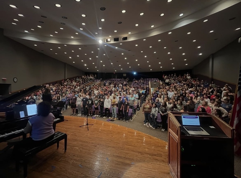 A large audience seated in a hall with a stage featuring two pianists, one seated, one standing, and a podium with laptops.