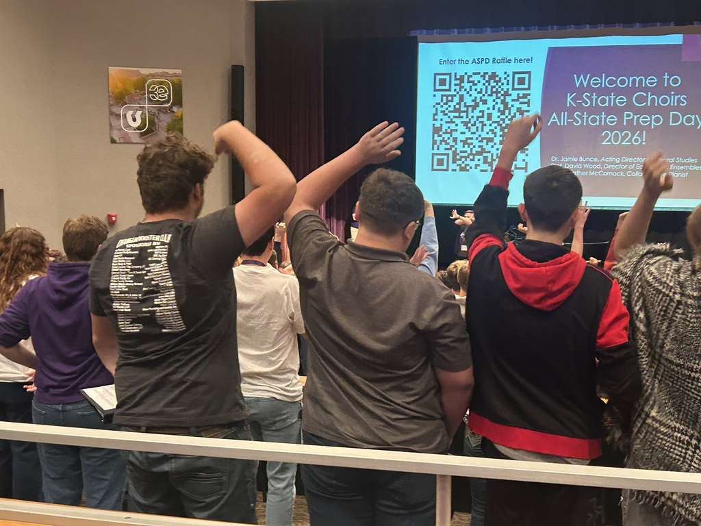 Several individuals are standing with raised hands in a classroom. A projector screen displays a presentation and a QR code.