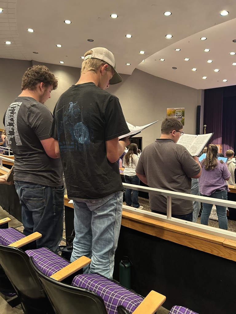 Four people stand in an auditorium, holding papers. One person wears a cap. Behind them, a row of chairs is visible.