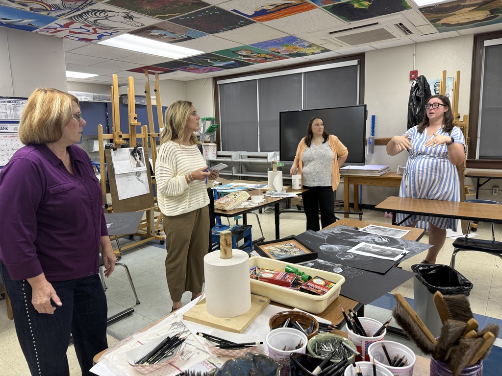 Four women in a room with tables and art supplies, one holding a clipboard.