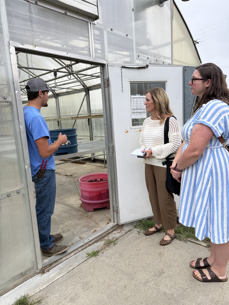 Three people stand in a greenhouse. A man in a hat and blue shirt, and two women, one with glasses.