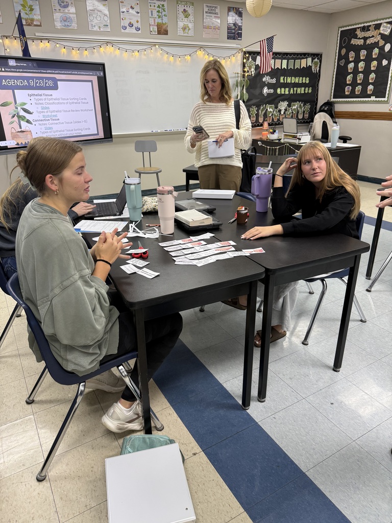 Two seated students focus on a table with objects. An instructor stands behind them, reading from a paper. Whiteboard, decorations, and a projector screen are in the background.