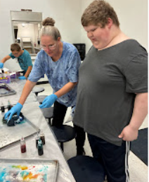 Two individuals wearing gloves work at a table. One stands, another bends over. A third person is behind them.