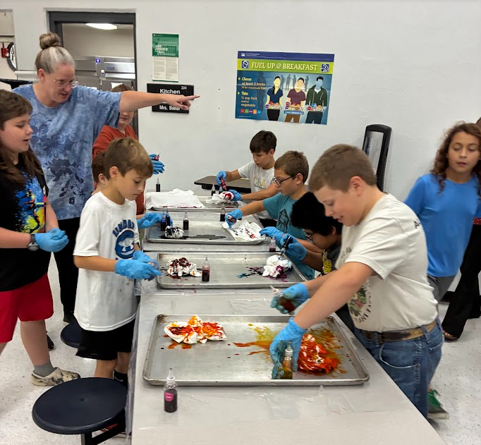 Children wearing gloves at a table mixing paint. Trays of paint and materials behind them.