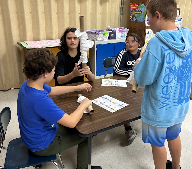 Three students in a classroom. One sits at a table, holding rolled paper, while two others stand, one wearing a blue hoodie.