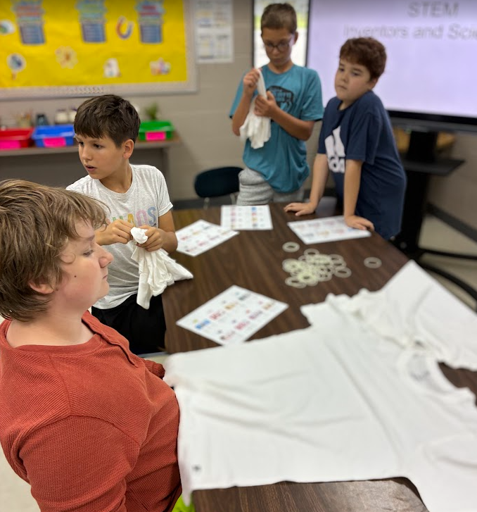 Four boys work together at a table, with one boy sitting and three boys standing, each holding a cloth.