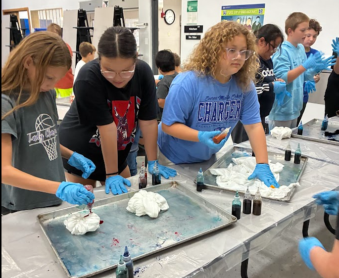 A group of children in gloves work on science projects in a classroom. They pour colored liquids on trays.
