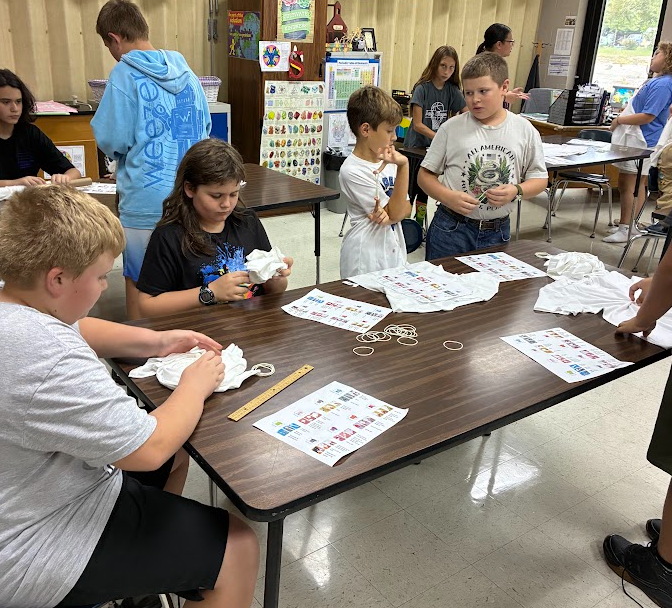 Several students in a classroom, seated around a table, working on a project with paper and string.