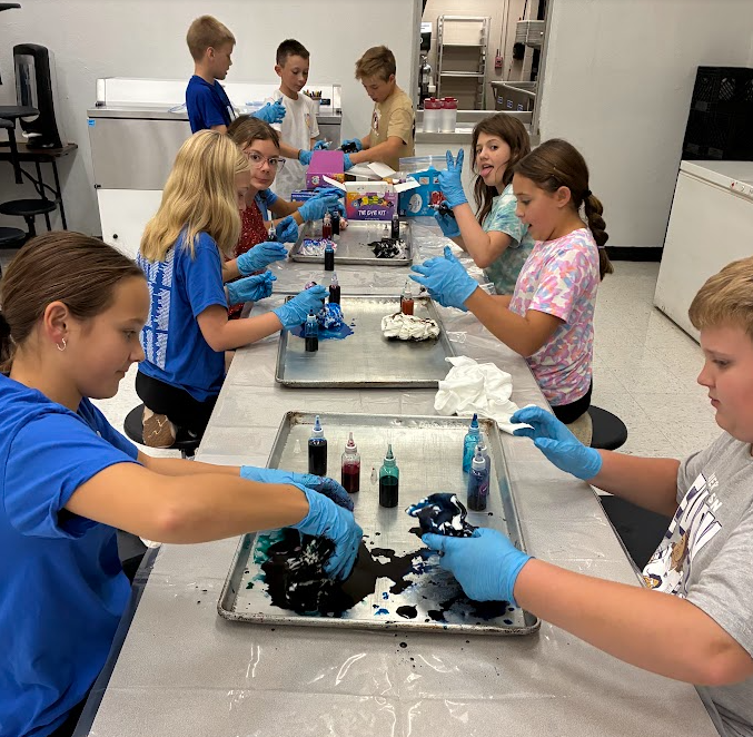Children wearing gloves at a table mixing paint. Trays of paint and materials behind them.