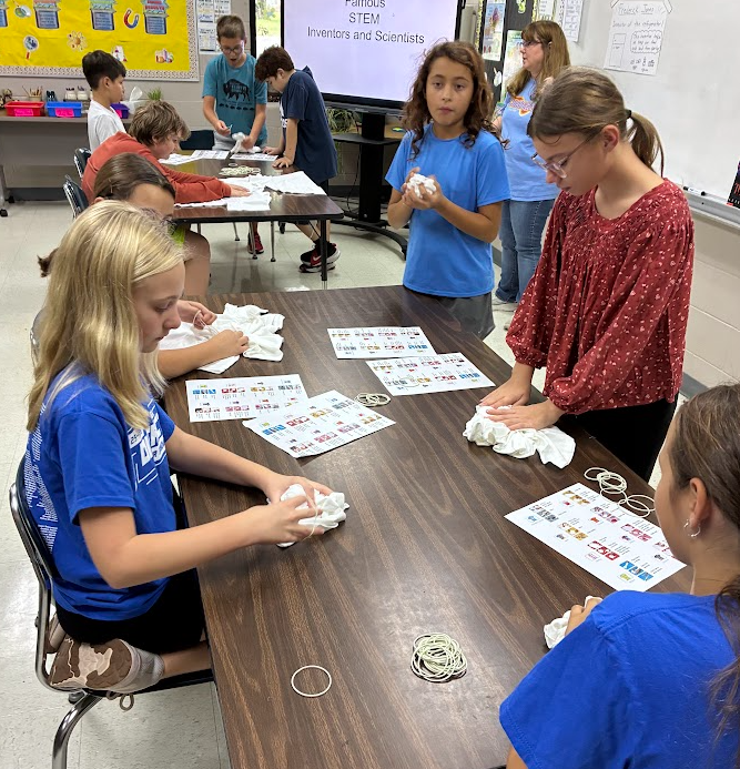 Several students sit around a table with science materials and papers. One holds a cloth.
