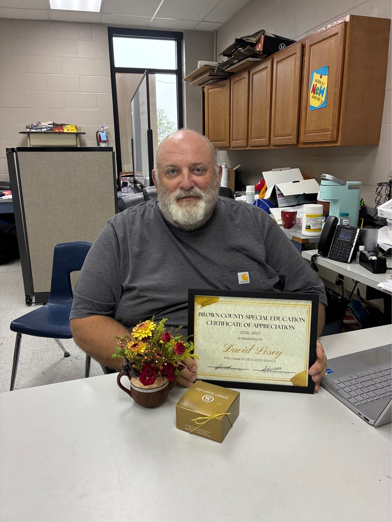 A man with a beard sits at a desk with a certificate, flowers, and a box.