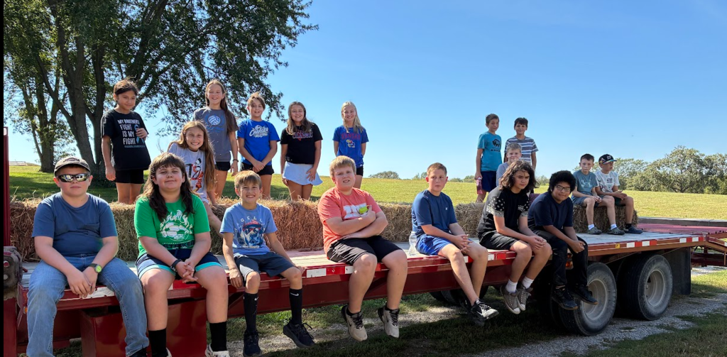 A group of children sit on the back of a truck on a sunny day in a grassy area.