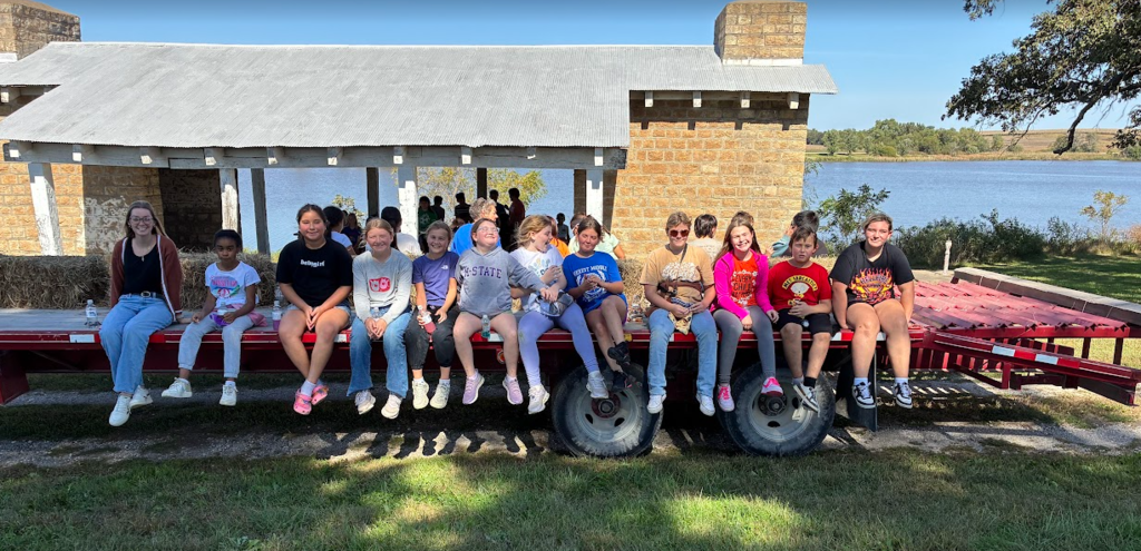 A group of children and adults sits on a flatbed trailer with a small house in the background.