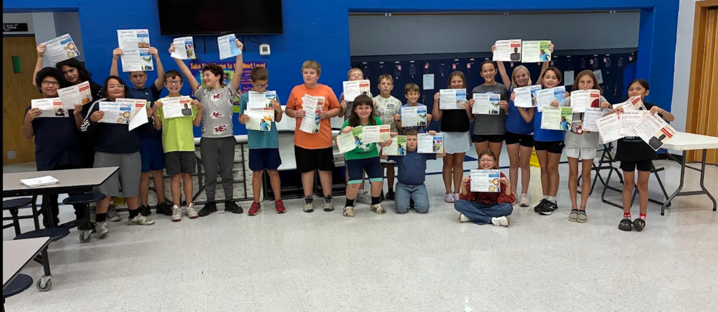 Children in a classroom hold certificates, standing and kneeling. Tables and a blue wall are in the background.