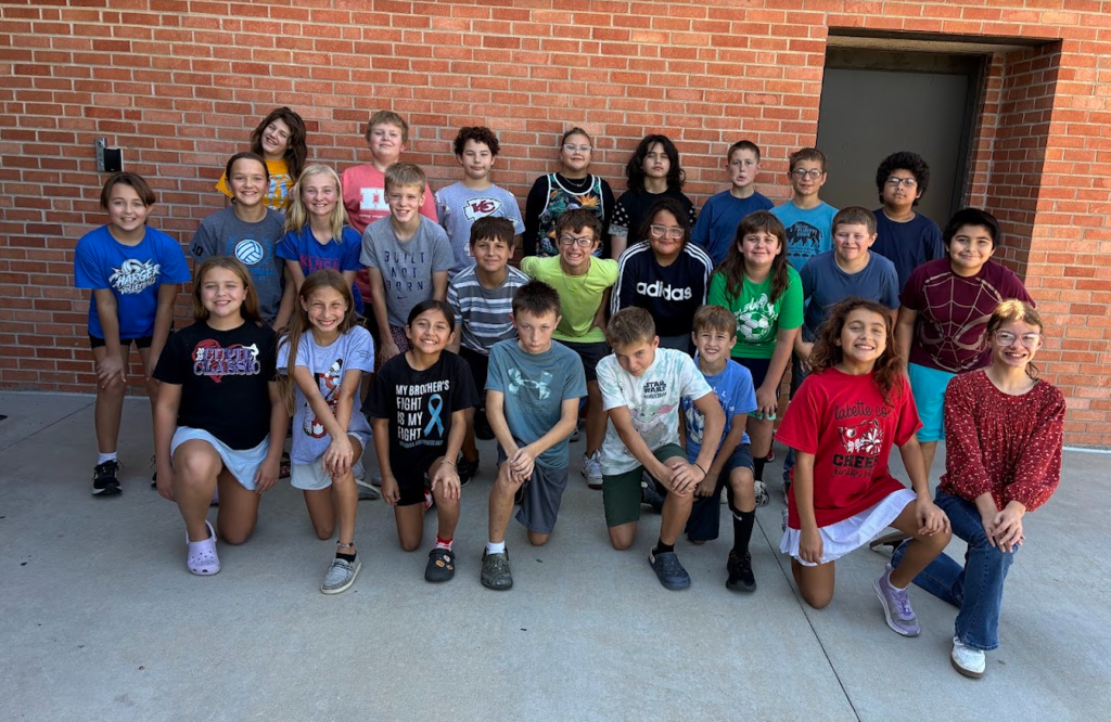 A group of young people in various shirts, sneakers, and shorts pose in front of a brick building.