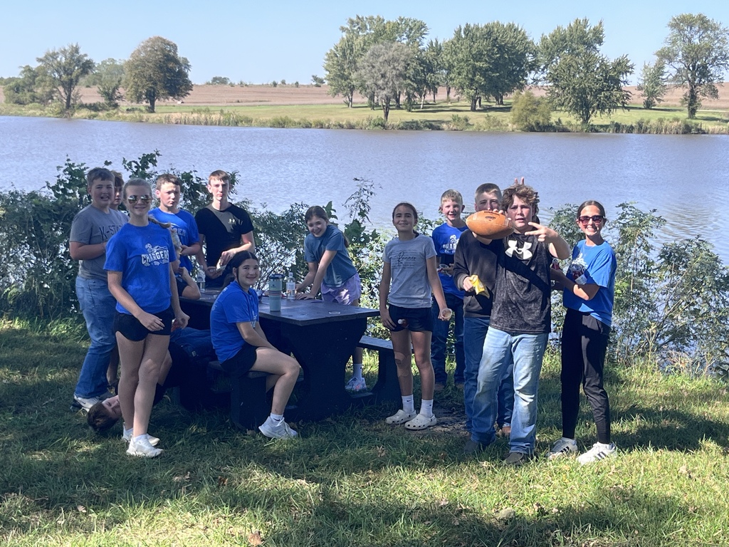 A group of people stands by a lake, some wearing blue shirts. They are posing with a football.