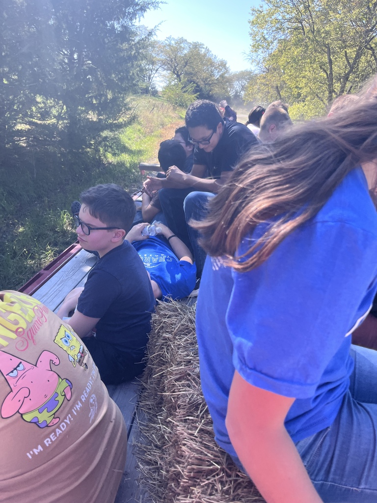 Several people sit on a bed of hay, possibly on a farm. A person wears glasses. A tree-filled background.