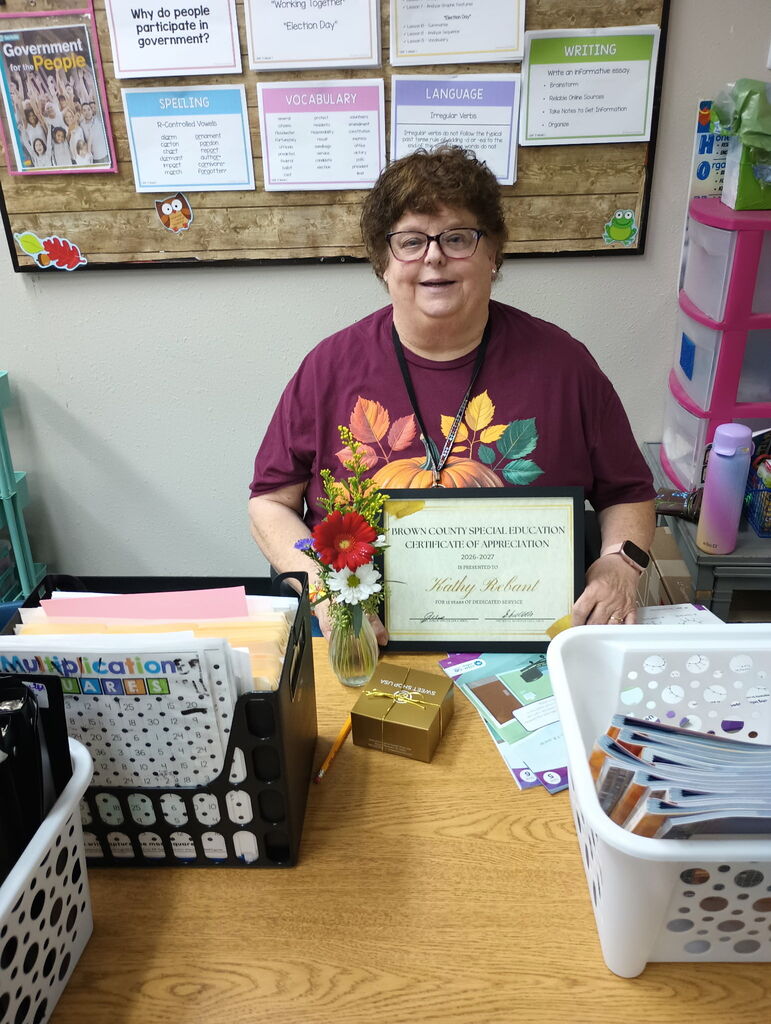 Person sitting at a desk, smiling, holding a framed award. Behind them, a bulletin board with papers attached.