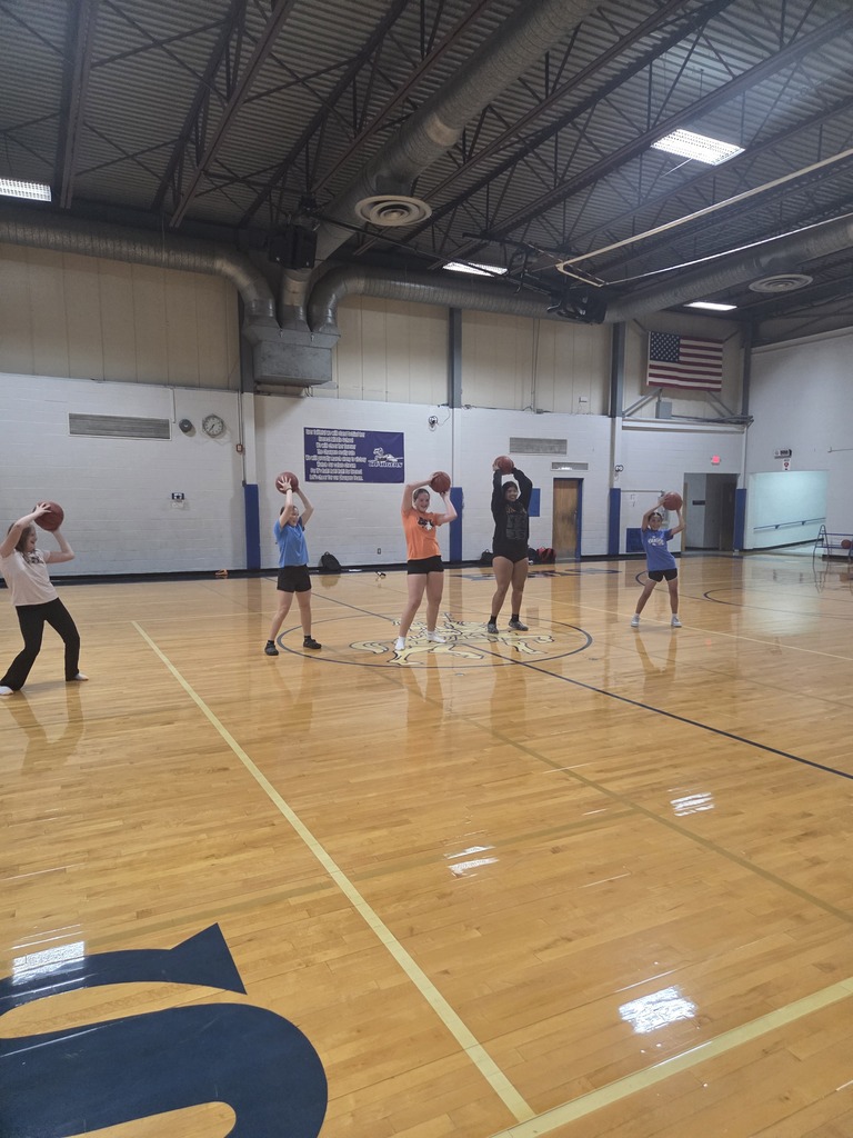 Several people play basketball in a gym. They hold balls and shoot hoops on a wooden floor.