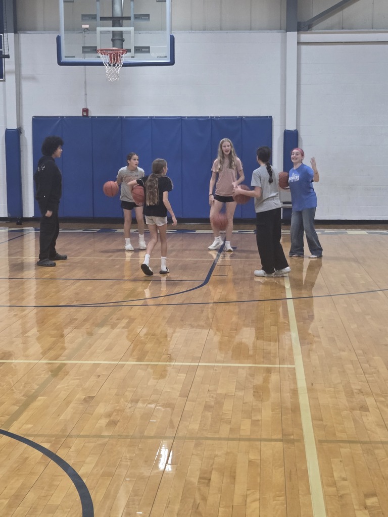 People wearing sports attire practice dribbling basketballs on an indoor court with blue walls and wooden flooring.