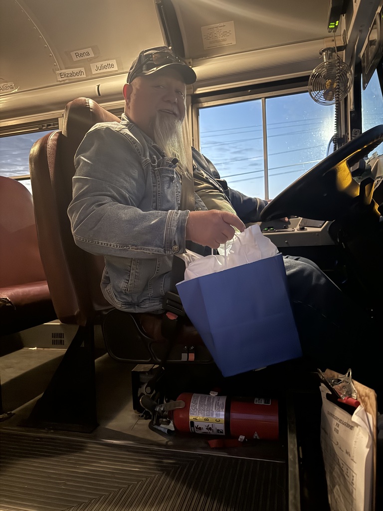 Man sitting in bus driver seat, holding gift bag and papers, wearing gray jacket and black hat.