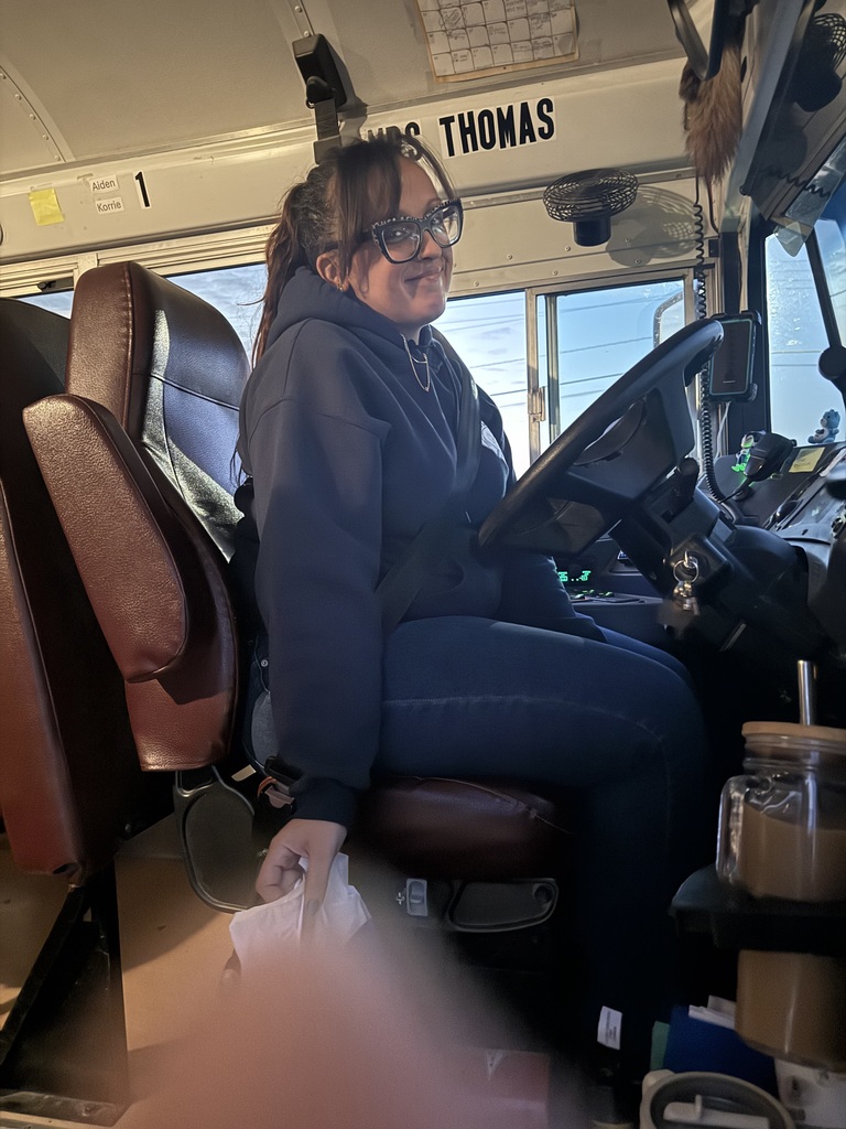 A woman sitting in a school bus, wearing glasses and a hoodie, holding a tissue.