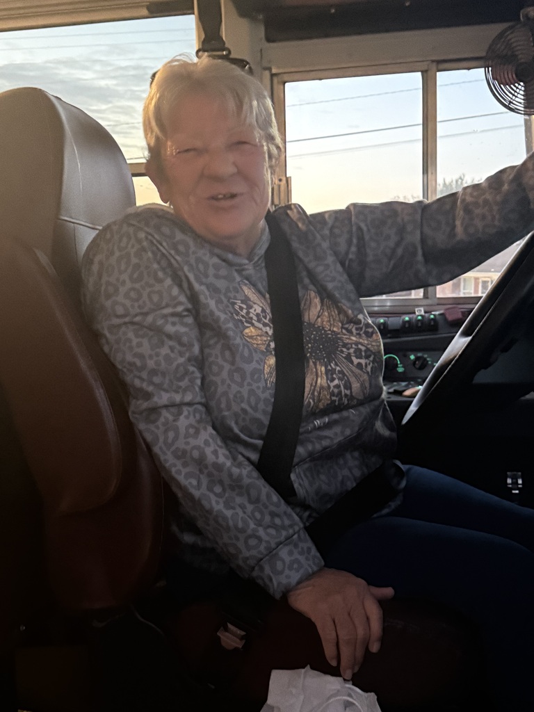 A woman wearing glasses sits on a school bus seat, smiling and looking forward with her hands on her lap.