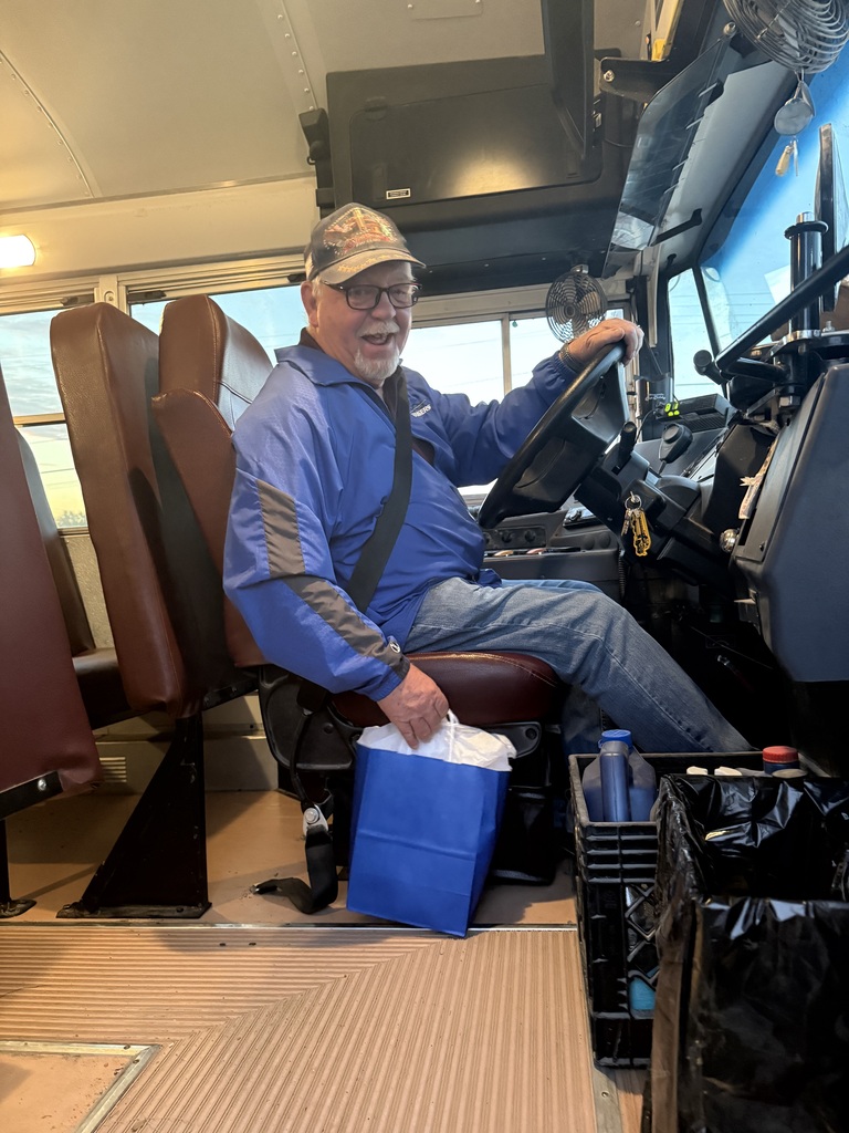 Man seated in bus driver's seat, holding steering wheel, wearing hat and jacket, with blue bag on lap.