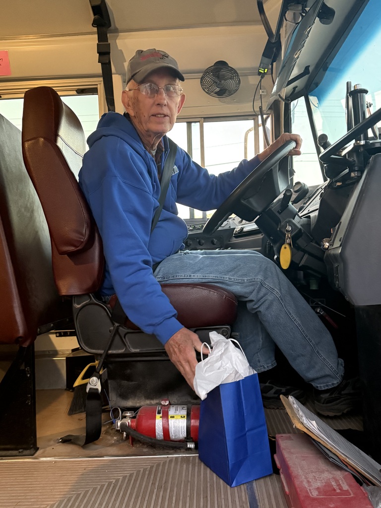 Elderly man sitting in a bus seat wearing a blue jacket, holding a bag and wearing a cap.