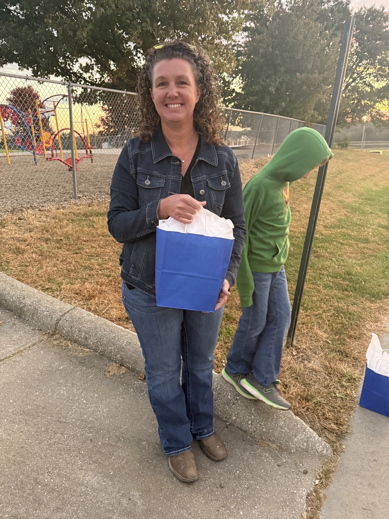 A woman with brown hair stands on a sidewalk holding a blue bag. A child stands nearby, wearing a green hoodie. Behind them, there is a grassy area and a fence.