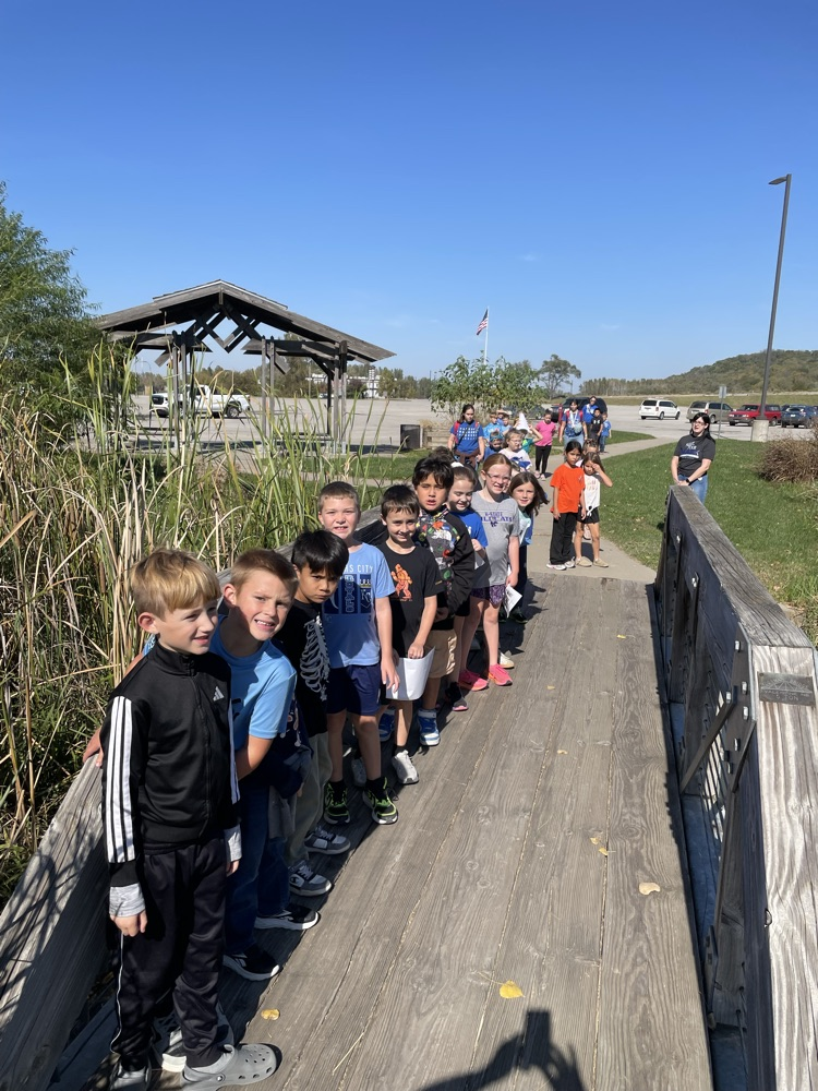 Children lined up on a wooden bridge, looking at tall grass and a gazebo in the background.