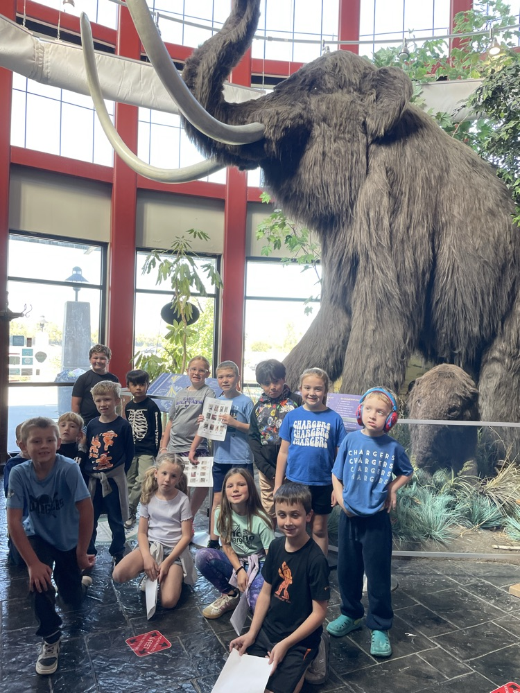 Group of young children standing and sitting in front of a large mammoth statue.