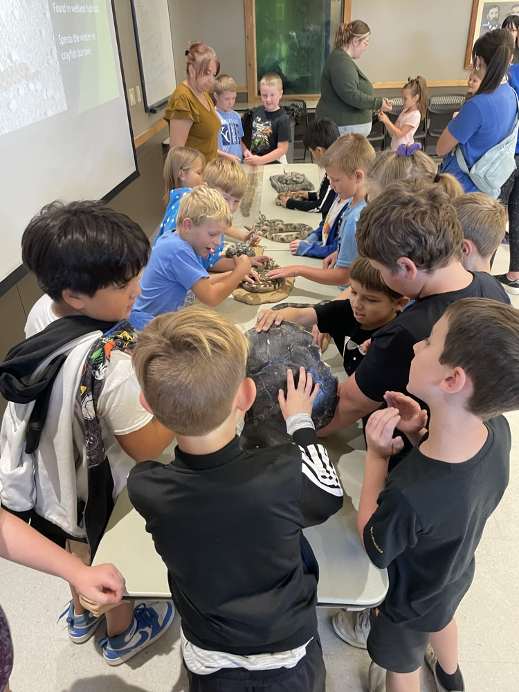 Children in a classroom, sitting at tables with hands on large gray rock, looking at each other.