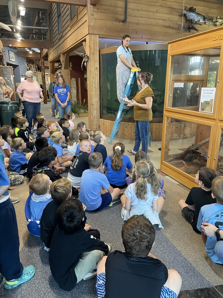Students sit on the floor around two people cleaning a glass tank with a ladder.