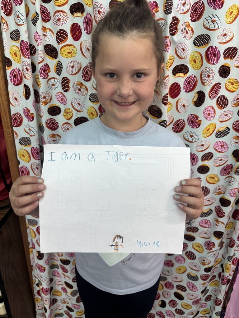 A young girl holds a sign that says "I am a Tiger" in front of a backdrop of colorful doughnuts.