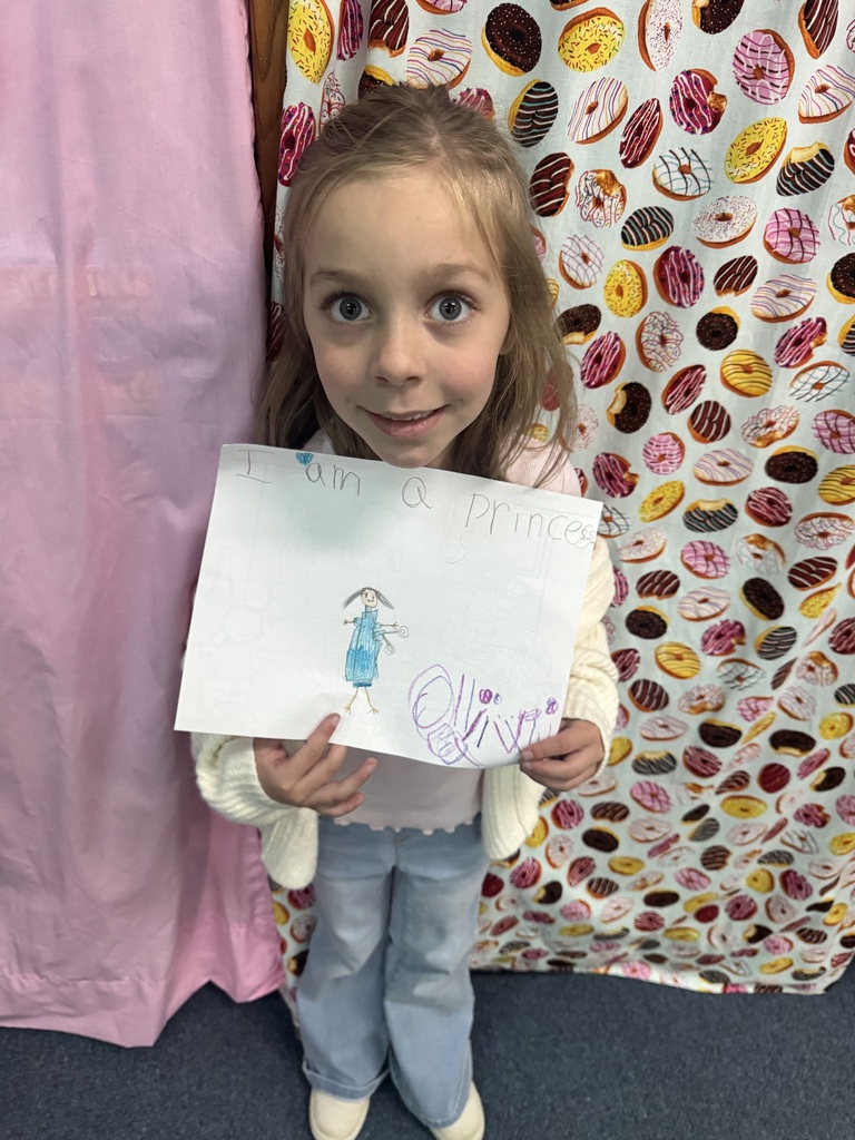 A girl in a white sweater holds a drawing of a girl in blue. She stands with pink curtains behind her.
