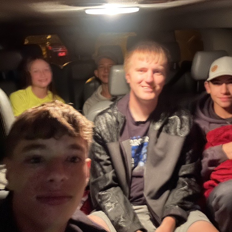 Four boys seated in a car, looking at the camera. The boy in front wears a black jacket.