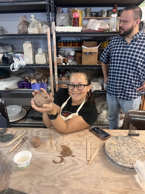 art teacher holding her piece of pottery, smiling. Man behind her in plaid looking off in the distance with hands on hips
