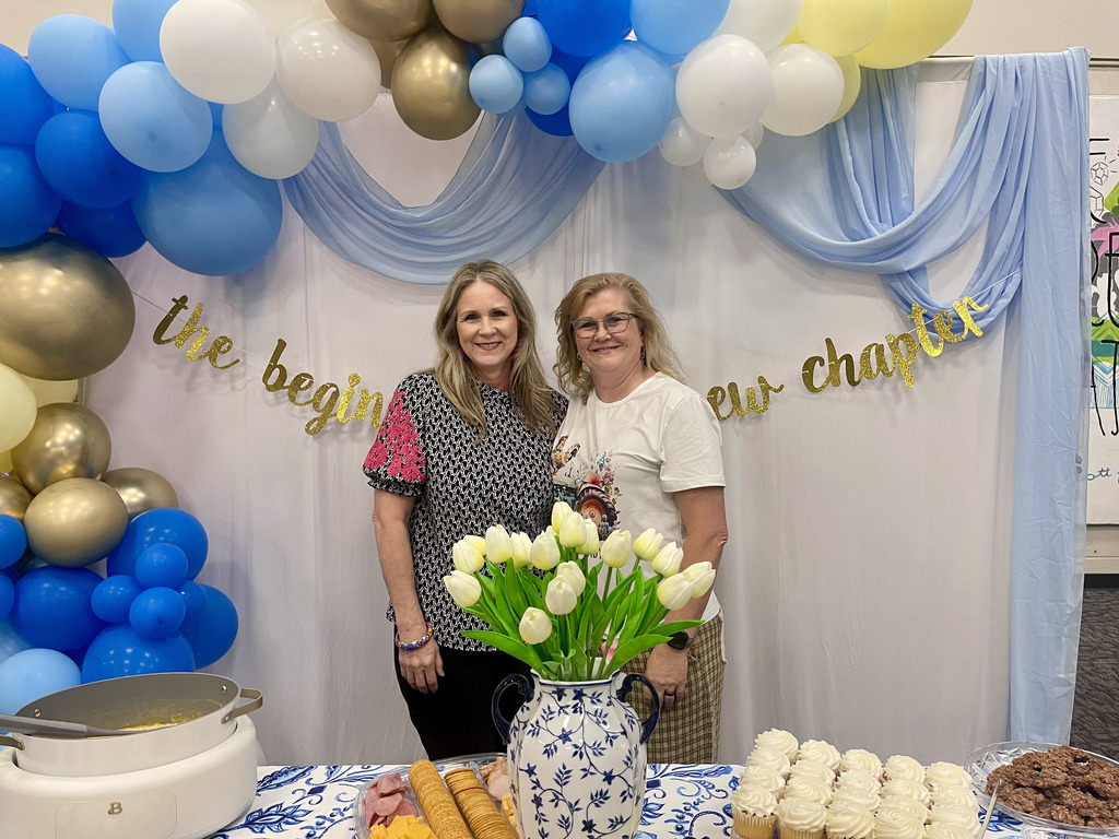 Kim and Stacy in front of decorated backdrop and in front of table of treats at retirement reception 