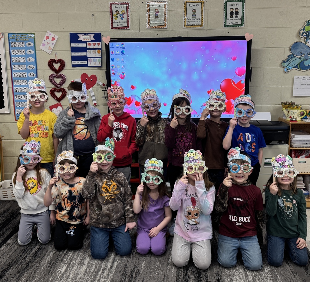 group of kindergarteners wearing 100 day smarters crown and classes posing in front of newline board in a classroom