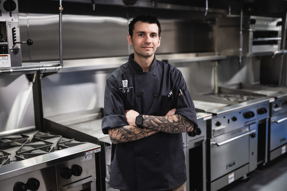 A Van Buren Tech alumni chef stands in a commercial kitchen with his arms crossed, wearing a black chef coat. Stainless steel stoves and kitchen equipment are visible behind him.