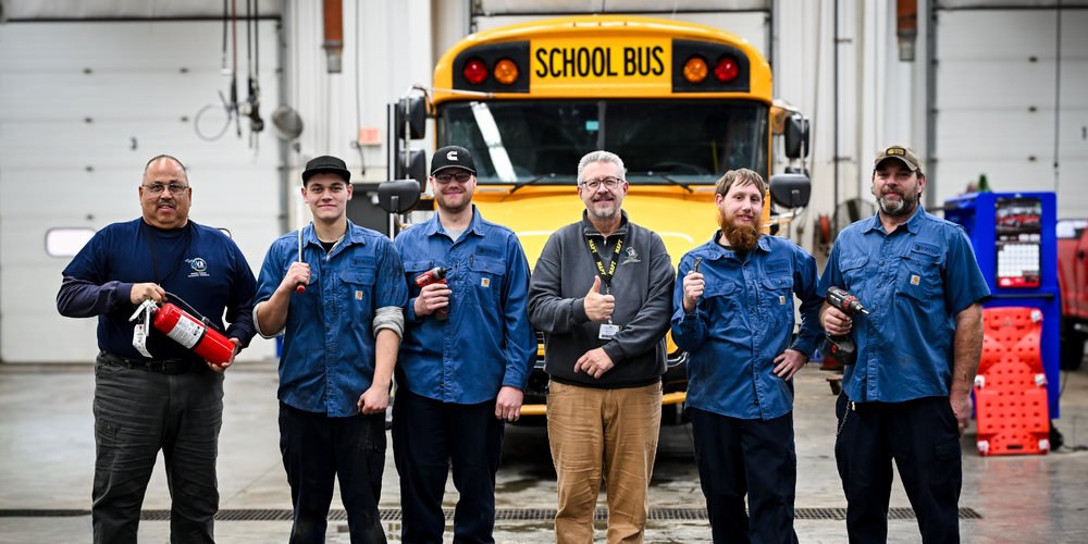 image of 6 unidentified men in blue shirts in front of a school bus holding up tools